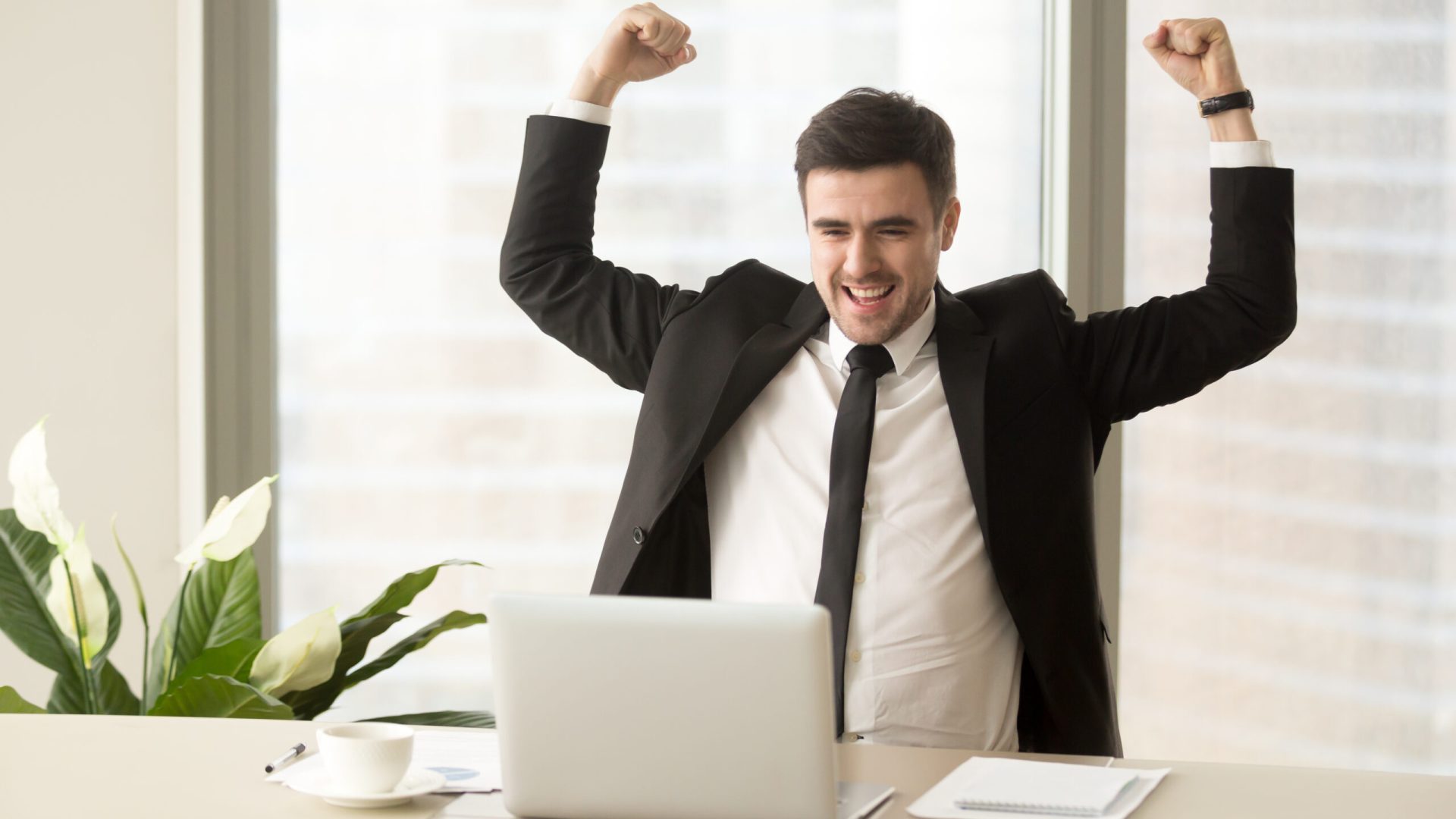 Millennial businessman raising hands and happily yelling when looking on laptop at desk. Business leader feeling positive because of successful deal confirmation, excited with great result in work
